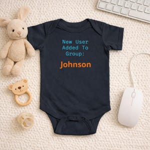 May include: Navy blue baby bodysuit with the text "New User Added To Group: Johnson" in blue and orange. A plush bunny, a wooden bear-shaped teether, and a rattle are next to the bodysuit.