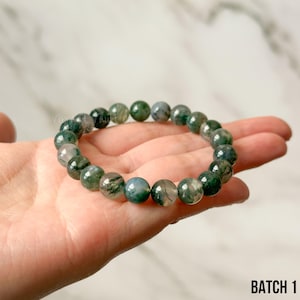 K&ouml;nnte beinhalten: Ein Armband mit runden Moosachat-Perlen in Gr&uuml;n- und Graut&ouml;nen. Die Steine werden in einer Hand gehalten, wodurch ihre nat&uuml;rlichen Muster hervorgehoben werden. Der Text "BATCH 1" ist unten rechts im Bild zu sehen.