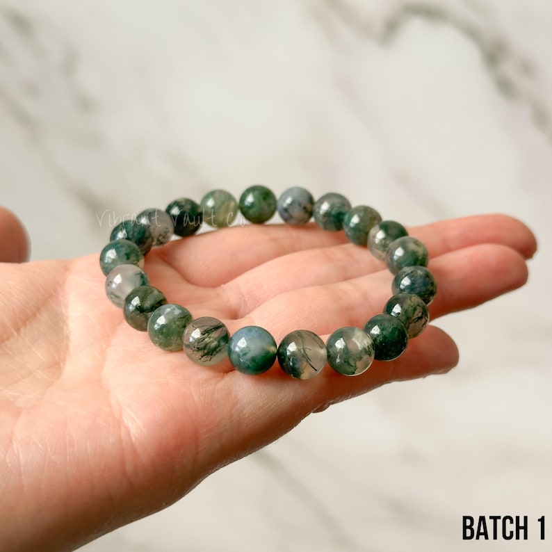 K&ouml;nnte beinhalten: Ein Armband mit runden Moosachat-Perlen in Gr&uuml;n- und Graut&ouml;nen. Die Steine werden in einer Hand gehalten, wodurch ihre nat&uuml;rlichen Muster hervorgehoben werden. Der Text "BATCH 1" ist unten rechts im Bild zu sehen.