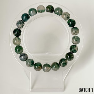 K&ouml;nnte beinhalten: Ein Armband mit runden Moosachat-Perlen in Gr&uuml;n- und Wei&szlig;t&ouml;nen. Das Armband wird auf einem transparenten, kreisf&ouml;rmigen St&auml;nder pr&auml;sentiert. Der Text "BATCH 1" ist unten rechts zu sehen.