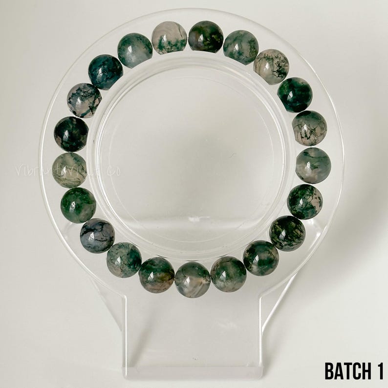 K&ouml;nnte beinhalten: Ein Armband mit runden Moosachat-Perlen in Gr&uuml;n- und Wei&szlig;t&ouml;nen. Das Armband wird auf einem transparenten, kreisf&ouml;rmigen St&auml;nder pr&auml;sentiert. Der Text "BATCH 1" ist unten rechts zu sehen.
