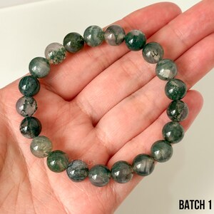 K&ouml;nnte beinhalten: Ein Armband aus runden Moosachat-Perlen in Gr&uuml;n- und Graut&ouml;nen. Die Perlen sind aufgereiht und bilden einen Kreis. Das Armband wird in einer Hand gehalten. Der Text "BATCH 1" ist unten rechts sichtbar.
