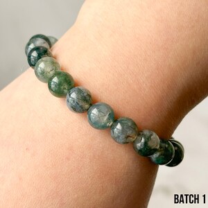 K&ouml;nnte beinhalten: Ein Armband mit runden Moosachat-Perlen in Gr&uuml;n- und Graut&ouml;nen. Das Armband wird am Handgelenk getragen. Der Text "BATCH 1" ist unten rechts sichtbar.