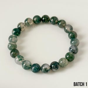 K&ouml;nnte beinhalten: Ein Armband mit runden Moosachat-Perlen. Die Perlen sind eine Mischung aus durchscheinendem Wei&szlig; und verschiedenen Gr&uuml;nt&ouml;nen. Das Armband wird auf einer wei&szlig;en Oberfl&auml;che pr&auml;sentiert. Der Text "BATCH 1" ist sichtbar.