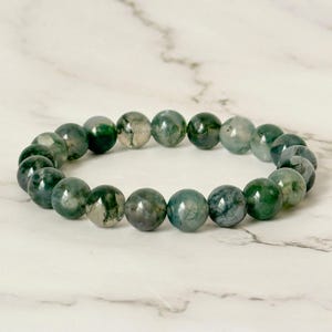 K&ouml;nnte beinhalten: Ein Armband mit runden Moosachat-Perlen in Gr&uuml;n- und Graut&ouml;nen. Die Perlen sind zu einem Kreis geformt. Das Armband liegt auf einer wei&szlig;en Marmoroberfl&auml;che.