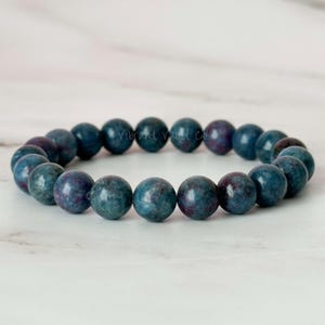 May include: A beaded bracelet featuring round, polished beads in shades of teal and purple. The bracelet is made of multiple beads strung together, creating a circular shape. The beads have a speckled appearance and a glossy finish.