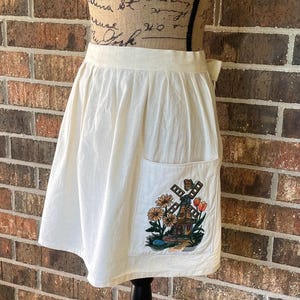 May include: Cream-colored apron with a pocket featuring an embroidered windmill scene with flowers. The apron is displayed on a mannequin and has a simple, classic design. The apron is made of a lightweight fabric.