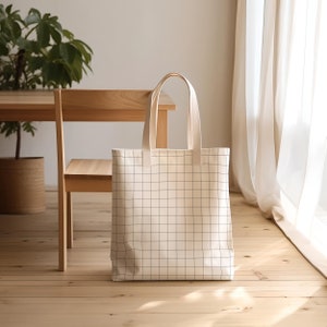 Puede incluir: Una bolsa de lona blanca con un patr&oacute;n de cuadr&iacute;cula negra. La bolsa tiene dos asas y est&aacute; sentada en un suelo de madera.