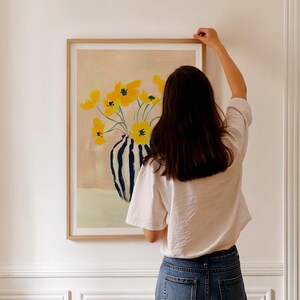 K&ouml;nnte beinhalten: Ein gerahmter Druck eines Gem&auml;ldes mit einem Strau&szlig; gelber Blumen in einer blau-wei&szlig; gestreiften Vase. Die Vase steht auf einem hellrosa Hintergrund.