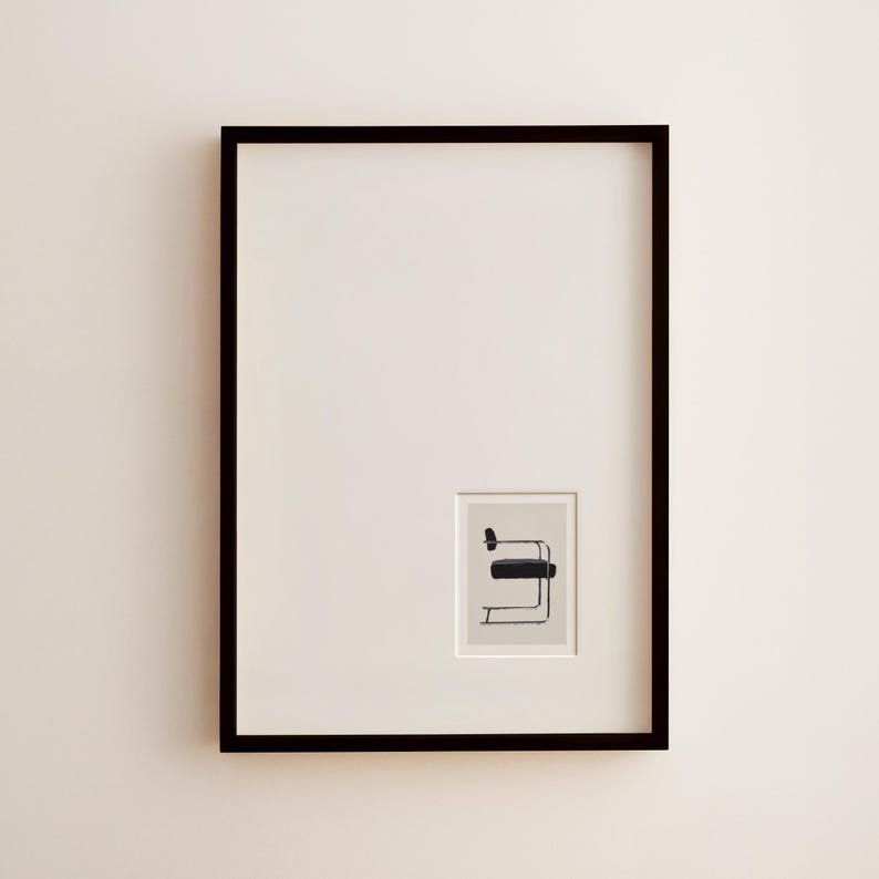 May include: A framed art print featuring a minimalist illustration of a black chair with a rectangular seat and curved armrest. The print is set against a cream-colored background within a black frame, creating a modern aesthetic.