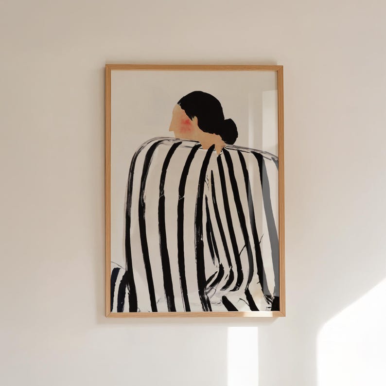 May include: Framed art print featuring a figure with a black bun and a striped black and white garment. The artwork is displayed in a light brown wooden frame, adding a touch of warmth to the minimalist design. The figure's face has a blush of pink.