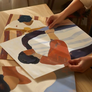 May include: A hand holds up a painting of two people in a stylized, abstract style. The painting is on a white background with blue and orange colors. The painting is on top of another painting with a brown and orange background.