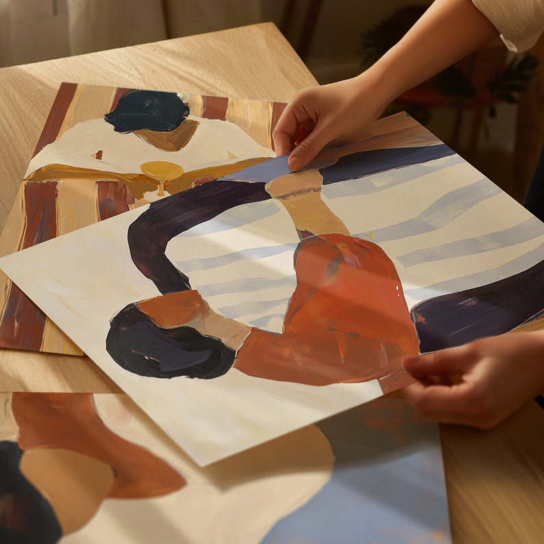 May include: A hand holds up a painting of two people in a stylized, abstract style. The painting is on a white background with blue and orange colors. The painting is on top of another painting with a brown and orange background.