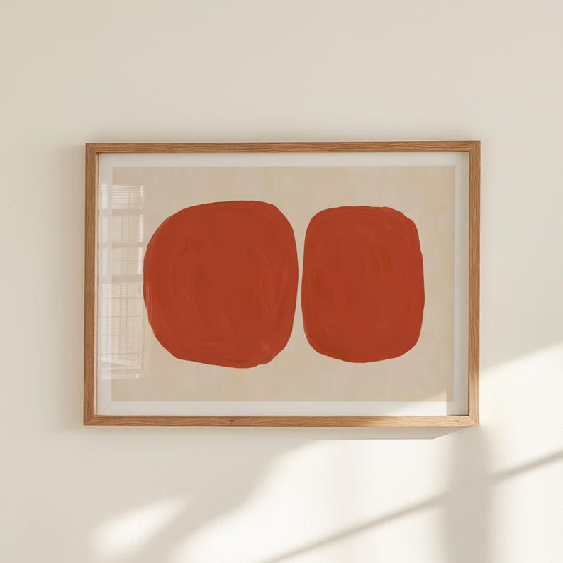 K&ouml;nnte beinhalten: Abstrakter Kunstdruck mit zwei gro&szlig;en, roten, organischen Formen auf cremefarbenem Hintergrund. Das Kunstwerk ist in einem hellbraunen Holzrahmen gerahmt. Eine moderne Wanddekoration, passend f&uuml;r verschiedene Einrichtungsstile.