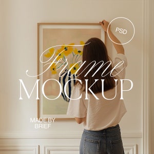 May include: A person hangs a framed print of a yellow flower painting on a white wall. The print is in a simple wooden frame. The text "Frame Mockup" and "Made by Brief" is visible below the print.