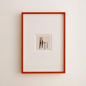 May include: A framed art print featuring a minimalist illustration of a chair. The chair has a light brown back and seat with black legs. The artwork is set within a white mat and a vibrant red frame.