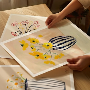 May include: Two prints of floral arrangements in vases. The top print features yellow flowers in a blue and white striped vase on a light pink background. The bottom print features red flowers in a black vase on a white background.