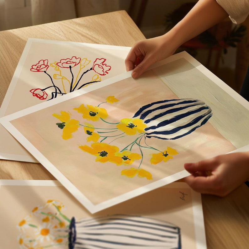 May include: Two prints of floral arrangements in vases. The top print features yellow flowers in a blue and white striped vase on a light pink background. The bottom print features red flowers in a black vase on a white background.