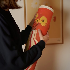 Puede incluir: Una persona sosteniendo un p&oacute;ster enrollado con fondo rojo y flores amarillas. El p&oacute;ster tiene un patr&oacute;n de rayas blancas en la mitad inferior.