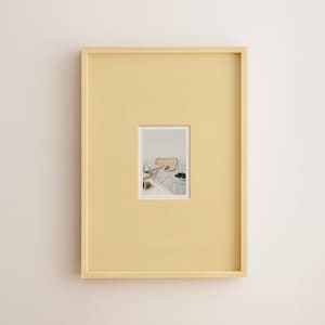 May include: A rectangular, pale yellow picture frame with a smaller, rectangular image inset. The image shows a table setting with a plate of food, a cup, and a bowl of dark berries. The frame is mounted on a white wall.