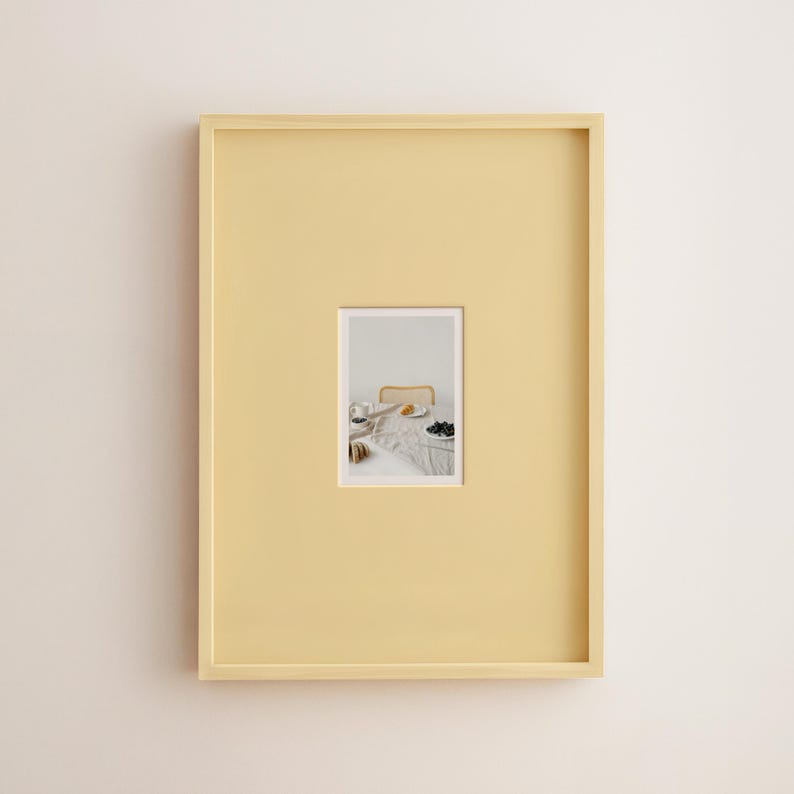 May include: A rectangular, pale yellow picture frame with a smaller, rectangular image inset. The image shows a table setting with a plate of food, a cup, and a bowl of dark berries. The frame is mounted on a white wall.