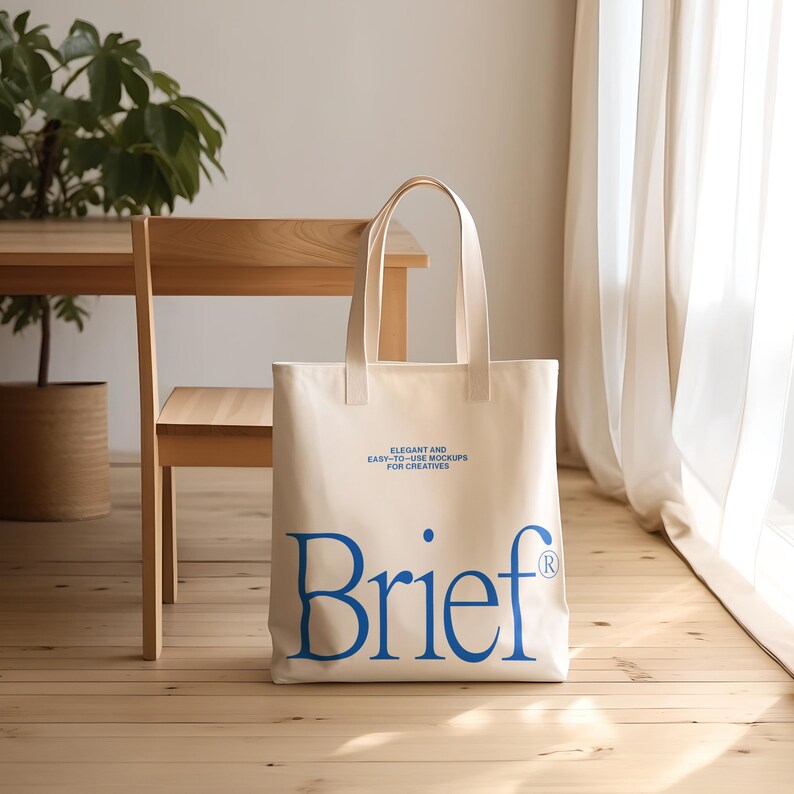 Puede incluir: Una bolsa de lona blanca con texto azul que dice "Elegant and Easy-to-Use Mockups for Creatives" y "Brief" con un s&iacute;mbolo de marca registrada. La bolsa est&aacute; sentada en un suelo de madera.