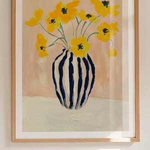 May include: A framed art print featuring a vase of yellow flowers. The vase has a blue and white striped pattern. The background is a soft pink, and the frame is a light wood color. The artwork is a floral still life.