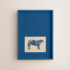 May include: A framed art print featuring a stylized blue cow with white spots on a beige background. The artwork is set within a vibrant blue frame, creating a striking contrast. The overall aesthetic is minimalist and modern, suitable for home decor.
