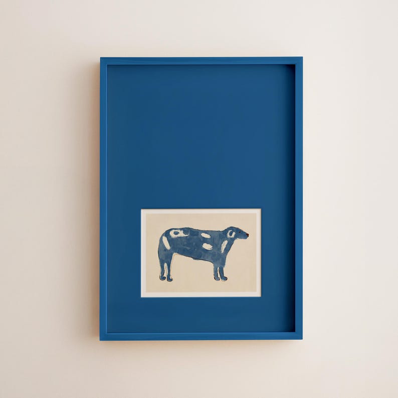 May include: A framed art print featuring a stylized blue cow with white spots on a beige background. The artwork is set within a vibrant blue frame, creating a striking contrast. The overall aesthetic is minimalist and modern, suitable for home decor.