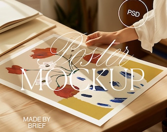 Puede incluir: Una maqueta de póster blanca con una ilustración floral de flores rojas y blancas en un jarrón azul y blanco. El póster está sobre una mesa de madera con el texto "Poster Mockup" y "Made by Brief" en el póster.