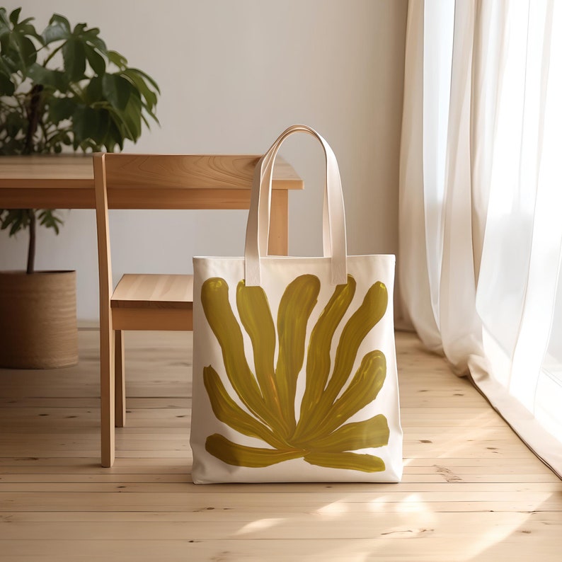 Puede incluir: Una bolsa de lona blanca con un gran dise&ntilde;o abstracto de hoja amarilla mostaza. La bolsa tiene dos asas largas.