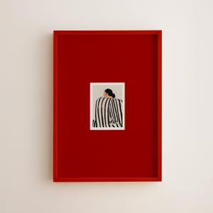 May include: A framed art print featuring a person in a black and white striped top, set against a neutral background. The print is encased in a vibrant red frame, creating a striking contrast against the white wall.