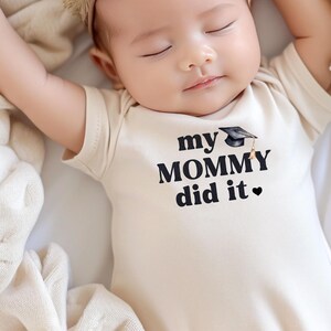 May include: A baby wearing a cream-colored onesie with the text "my MOMMY did it" and a black graduation cap graphic. The baby is sleeping with arms outstretched.