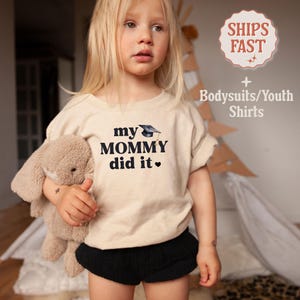 May include: A toddler wearing a beige t-shirt with the text "my mommy did it" and a graduation cap graphic. The shirt is paired with black shorts.