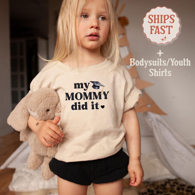 May include: A toddler wearing a beige t-shirt with the text "my mommy did it" and a graduation cap graphic. The shirt is paired with black shorts.