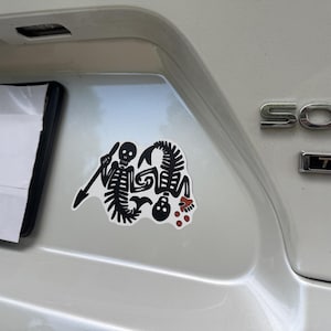May include: A black and white sticker featuring a skeleton with a spear and a stylized skeleton-like creature. The sticker has red accents and is affixed to the rear of a white vehicle.