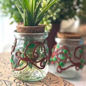 May include: Two glass jars with cork lids decorated with brown and green clay vines. The jars contain plants with roots visible in the water.
