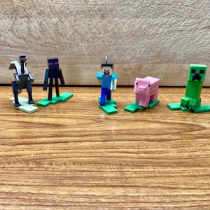 May include: Five Minecraft character figurines on green bases. The characters are a black and purple figure, a blue figure with a pickaxe, a pink pig, a green creeper, and a gray and silver figure.