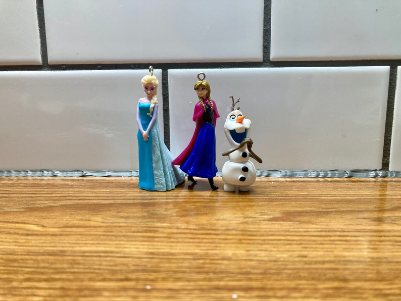 Puede incluir: Tres peque&ntilde;as figuras de pl&aacute;stico de personajes de la pel&iacute;cula Frozen. Las figuras son Elsa, Anna y Olaf. Elsa lleva un vestido azul claro con una capa larga y fluida. Anna lleva un vestido azul oscuro con una capa roja. Olaf es un mu&ntilde;eco de nieve con una nariz de zanahoria y una gran sonrisa.