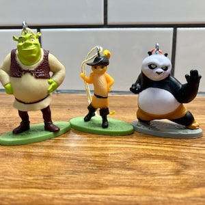 Pode incluir: Três ornamentos de personagens de desenhos animados verdes, amarelos e pretos. Shrek, o Gato de Botas e Po de Kung Fu Panda.