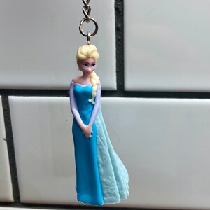 Puede incluir: Una figurita de pl&aacute;stico azul y blanco de una mujer con cabello rubio que lleva un vestido azul y una capa blanca. La figurita est&aacute; unida a un llavero plateado.