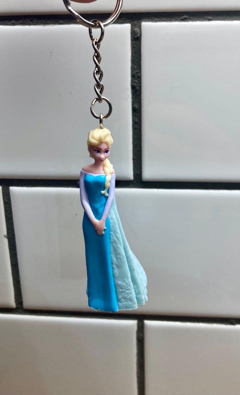 Puede incluir: Una figurita de pl&aacute;stico azul y blanco de una mujer con cabello rubio que lleva un vestido azul y una capa blanca. La figurita est&aacute; unida a un llavero plateado.