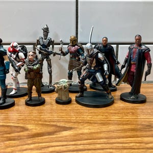 May include: A collection of eight Star Wars action figures, including a Mandalorian, a Stormtrooper, a Rebel pilot, and a Jedi. The figures are all standing on small black bases.