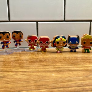 Puede incluir: Un conjunto de siete figuras de superhéroes, incluyendo Superman, Batman, Wonder Woman, Flash y Aquaman. Las figuras están todas en un estilo pop art y están de pie sobre una superficie de madera.
