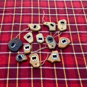 May include: A collection of wooden bottle openers in various shapes, including rectangular, teardrop, and organic forms. Each opener has a silver metal bottle cap remover and is attached to a natural twine string. The openers are displayed on a red and yellow plaid fabric.