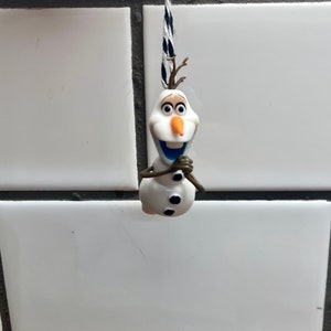 Puede incluir: Una figurita de mu&ntilde;eco de nieve blanco con ojos y botones negros, una nariz naranja y una sonrisa azul, colgada de una cuerda negra y blanca.