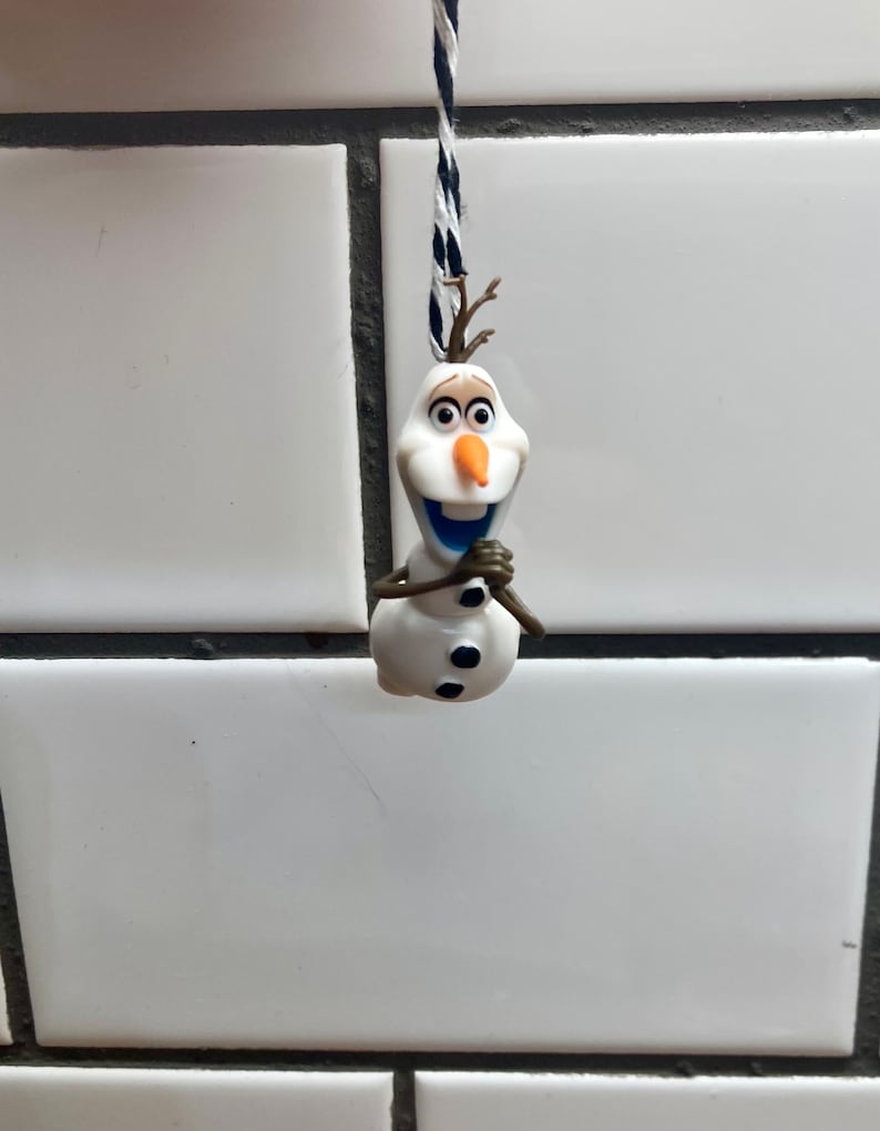 Puede incluir: Una figurita de mu&ntilde;eco de nieve blanco con ojos y botones negros, una nariz naranja y una sonrisa azul, colgada de una cuerda negra y blanca.