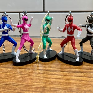 May include: Five Power Rangers action figures, each with a different color and design. The figures are posed in fighting stances and are attached to strings. The figures are on black bases with the words "Power Rangers" printed on them.