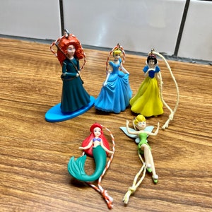 Puede incluir: Cinco adornos navideños de princesas Disney, incluyendo Ariel la sirena, Campanita, Cenicienta, Blancanieves y Mérida. Los adornos están hechos de plástico y tienen un acabado brillante.