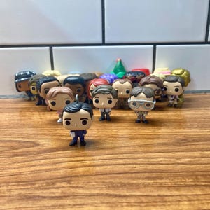 May include: A collection of 17 small, colorful, plastic figurines of characters from the TV show "The Office". The figurines are arranged in a semi-circle on a wooden surface.